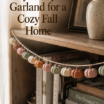 A homemade mini pumpkin garland of about eighteen small handmade air dry clay pumpkins in muted sage, cream, dusty rose, and warm cinnamon, strung on jute twine and draped naturally along a wooden bookshelf edge with books and dried grasses nearby.