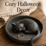 Small handmade round clay trinket dish with a sleeping curled charcoal-grey cat sculpted into the center and tail draped along the rim, holding a delicate gold ring, sitting on a wooden dresser with a folded linen napkin nearby and natural window light.