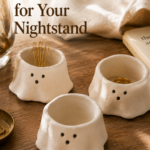 Three small chubby handmade white clay ghost pinch pots with off-center dot eyes and tiny mouths, one holding gold hair pins, one holding a small ring, arranged on a wooden nightstand with warm morning window light.