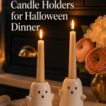 Two chubby handmade white clay ghost candle holders with off-center dot eyes and tiny mouths, each holding a lit cream taper candle, sitting on a wooden dining table at dusk with warm candlelight flickering across the surface.