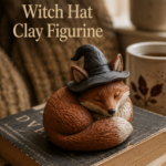 Small handmade curled sleeping fox clay figurine painted warm cinnamon with a cream chest, wearing a floppy soft charcoal witch hat perched between the ears, sitting on a stack of used paperback books on a wooden side table with warm window light.