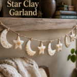 Handmade garland of small clay crescent moons and puffy stars painted cream with hand-brushed warm gold accents, strung on jute twine and draped naturally along a wooden shelf, with a soft folded blanket on a chair below and natural window light.