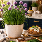Lush chive plant with tall slender green stalks and rounded purple pom-pom flowers in a cream ceramic pot on a white quartz countertop, dark navy cabinetry blurred behind, freshly chopped chives on a wooden cutting board beside a rustic baked potato topped with a dollop of sour cream, brushed gold serving spoon nearby, warm natural light.