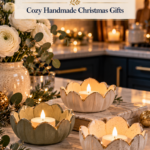 Three petal-shaped air dry clay candle holders in cream, speckled, and sage with gold-painted edges, holding glowing tealights on a marble kitchen island with white ranunculus, eucalyptus, and evergreen. The text reads "Air Dry Clay Tealight Holders, Cozy Handmade Christmas Gifts" and "Tap to Make Your Own."