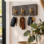 A DIY air dry clay key holder with a dark stone-look finish and three brass hooks holding car keys, mounted on an entryway wall above a wood console with a speckled bowl and eucalyptus. The text reads "DIY Clay Key Holder, Never Lose Your Keys Again" and "Save for Gift Ideas."