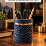 A navy air dry clay pen cup with a braided rope texture and a gold band, holding black and gold pens and a pencil on a wooden desk with a brass lamp, a succulent, and leather notebooks. The text reads "Textured Clay Desk Pen Cup, Minimal Handmade Gifts for Men" and "Tap to Make Yours."