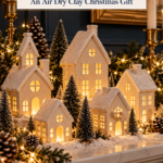 A mantel display of white air dry clay Christmas village houses glowing from inside, surrounded by bottle brush trees, pinecones, fairy lights, and brass candlesticks. The text reads "Glowing Clay Christmas Village, An Air Dry Clay Christmas Gift" and "Tap to Make Yours."