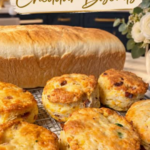 Golden jalapeño bacon cheddar biscuits cooling on a wire rack in front of a homemade loaf of bread, with visible bacon pieces and green jalapeño, a white ranunculus arrangement, and a navy kitchen with a gold faucet behind