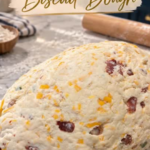A large mound of soft biscuit dough studded with shredded cheddar, bacon, and jalapeño on a floured marble counter, with a wooden rolling pin, a bowl of flour, and a striped towel nearby.