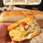 A hand holding a split homemade cheddar tea biscuit showing flaky layers, melted cheddar, and a piece of bacon, with loaves of homemade bread and a wire cooling rack blurred in a navy and gold kitchen behind.
