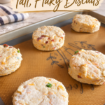 Five unbaked round cheddar biscuits speckled with bacon and jalapeño on a gold silicone baking mat in a sheet pan, freshly cut with straight sides, with a striped kitchen towel and flowers on a marble counter behind.