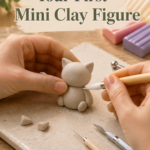Close-up of hands shaping a small gray clay cat figure on a ceramic tile, using a ball-tip sculpting tool, with pastel polymer clay bars and additional tools nearby. Board: your main clay/craft board