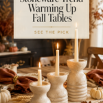 Three ribbed cream stoneware pillar candle holders in graduating heights clustered down the center of a rustic oak dining table with an oatmeal linen runner, rust-colored napkins, brushed gold napkin rings, dried wheat, small pumpkins, and lit tapered candles casting warm candlelight for a cozy autumn tablescape.