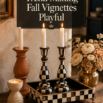 Two black-and-cream checkerboard patterned pillar candlestick holders with brushed gold accent bands sit on a matching checkered tray holding lit cream tapered candles, styled beside a plain unglazed terracotta pillar holder and a ribbed cream stoneware votive on a weathered oak console table with ivory ranunculus and dried cream hydrangea for a playful yet elevated fall vignette