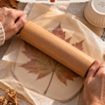 Hands pressing a wooden rolling pin firmly over parchment paper covering an air dry clay slab with a maple leaf pressed into it, showing the technique for getting crisp leaf vein detail in DIY clay leaf bowls.