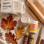 Overhead flat-lay of air dry clay leaf bowl supplies on cream linen — white clay block, wooden rolling pin, parchment paper, small ramekin, paring knife, sandpaper, Mod Podge, rust and gold acrylic paint bottles, and real fall maple and oak leaves.