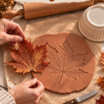 Overhead in-progress shot of a terracotta air dry clay slab with a maple leaf freshly peeled away, revealing a crisp leaf imprint. A wooden rolling pin, paring knife, and upside-down ceramic ramekin sit nearby on a cream linen surface for shaping the clay leaf bowl.