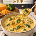 Brushed gold spoon lifting a bite of creamy keto copycat Panera broccoli cheddar soup with visible cheese pull and bright green broccoli from a rustic cream stoneware bowl, styled on white quartz counter with linen napkin, block of cheddar, and dark navy cabinetry in the background.
