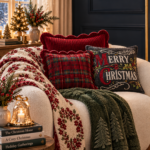 Cream boucle sofa layered with cozy Christmas textiles against a dark navy accent wall. Pillows include a scalloped red velvet pillow, a red and green plaid pillow, and a chalkboard "Merry Christmas" embroidered pillow. A floral wreath print quilt drapes across one arm, a deep green evergreen tree textured throw drapes across the other. Small side table with a bell candle and stack of Christmas books beside a mini lit tabletop tree, brass floor lamp, and framed "A Cozier Kinder Brighter Christmas" print. Text overlay reads "Layer Your Sofa With Cozy Christmas Warmth".