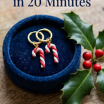 Gold hoop earrings with small red and white polymer clay candy cane charms on a navy velvet ring dish beside a sprig of holly with red berries.