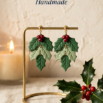 Layered green polymer clay holly leaf drop earrings with red berry clusters on gold studs, displayed on a gold stand beside a candle and fresh holly.
