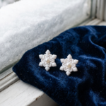 Pair of white polymer clay snowflake stud earrings with pearl centers resting on navy velvet on a snowy windowsill.