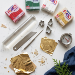 Flat lay of polymer clay supplies on white marble, including red, green, white, and gold Fimo Soft blocks, an acrylic roller, a blade, mini cutters, gold leaf, earring hooks, jump rings, and an oven thermometer.