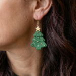 Close-up of a woman with long dark hair wearing a green polymer clay Christmas tree earring with tiny gold dots and a gold star on a gold hook.