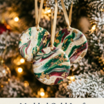 Three round marbled polymer clay ornaments in white, emerald green, and burgundy swirls with gold leaf flakes, hanging from gold ribbons on a flocked Christmas tree with warm lights.