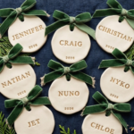 Eight cream polymer clay ornaments stamped with family names and 2026 in gold letters, tied with green velvet bows on a navy linen runner with cedar sprigs.