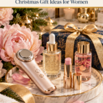 A rose-gold red light skincare wand glowing inside an open luxury gift box with cream tissue paper on a marble vanity, surrounded by gold ribbon, peony petals, and a serum bottle.