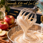 A mint blue stand mixer lifting its whisk out of a stainless bowl of thick, glossy, cream-colored spiced apple cider marshmallow fluff flecked with cinnamon, with a ribbon of fluff falling back into the bowl, beside red apples, apple slices, cinnamon sticks and a bowl of cinnamon in a navy kitchen.