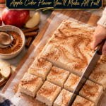 A cinnamon-swirled slab of homemade low carb apple cider marshmallows on parchment over a wooden cutting board, being sliced into neat squares with a large knife, beside a bowl of cinnamon with a sifter, red apples, apple slices, cinnamon sticks and a blue plaid towel.