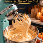 A mint blue stand mixer lifting its whisk out of a bowl of thick, glossy, golden pumpkin spice latte marshmallow fluff flecked with cinnamon, with a ribbon of fluff falling back into the stainless bowl, in a cozy navy kitchen with mini pumpkins, a candle and cinnamon sticks.