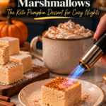 A hand using a kitchen torch to toast the top of a sugar-free pumpkin spice marshmallow until golden and caramelized on a speckled plate, with a mug of keto hot chocolate topped with whipped cream, a stack of cinnamon-dusted marshmallow squares on a wood board, cinnamon sticks and an orange pumpkin behind.